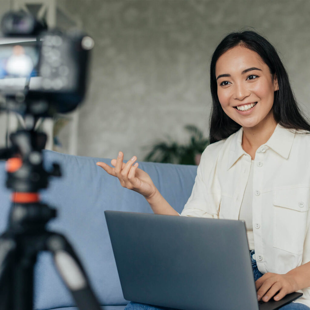 A woman sits and talks in front of a DLSR camera, carrying a laptop on her lap